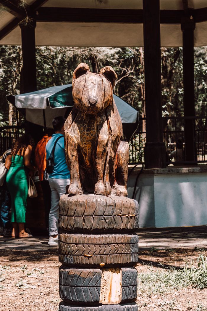 digital Wooden bear sculpture atop stacked tires in a sunny park area.